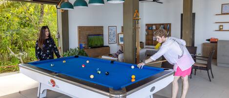 An indoor pool table with players, surrounded by a wooden ceiling, a television, and a bookshelf.