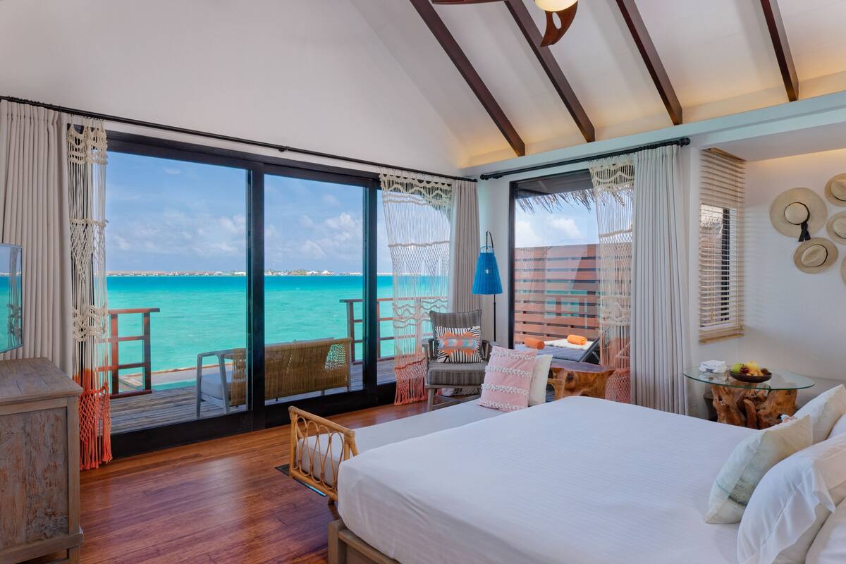 A bedroom with a large bed, a wooden floor, a view of the ocean, a balcony with wicker furniture, and a tropical decor theme.