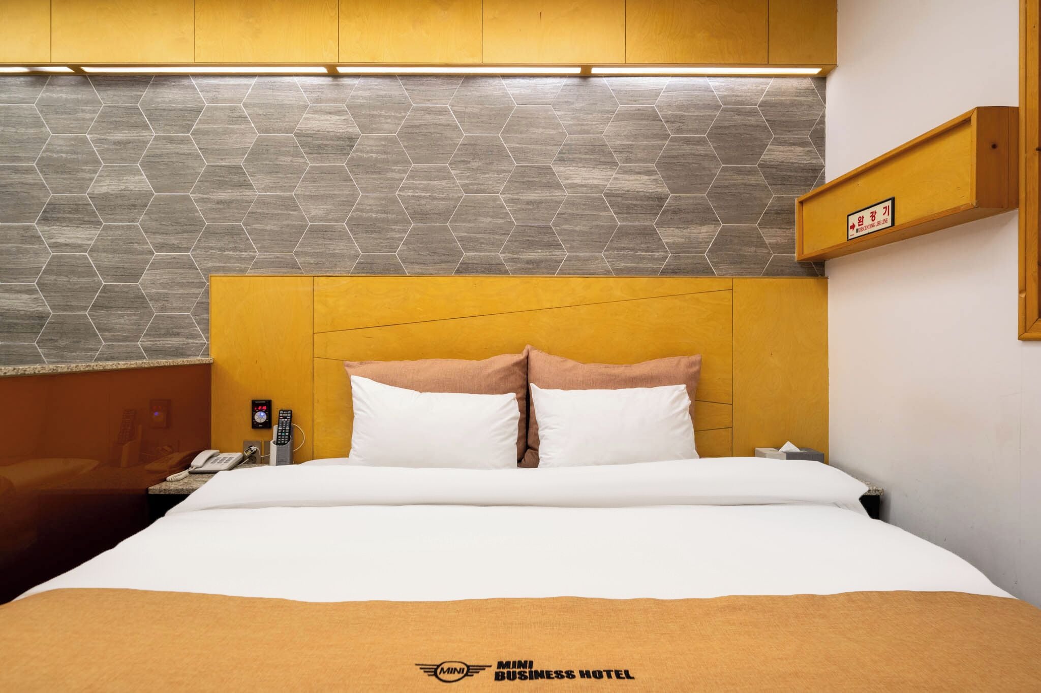 Una habitación de hotel con una cama, una mesita de noche con un teléfono y un control remoto, un estante en la pared con un letrero, y una pared con un patrón hexagonal.