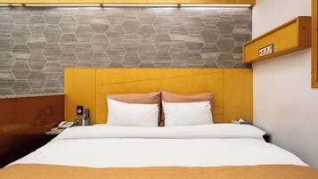 Una habitación de hotel con una cama, una mesita de noche con un teléfono y un control remoto, un estante en la pared con un letrero, y una pared con un patrón hexagonal.
