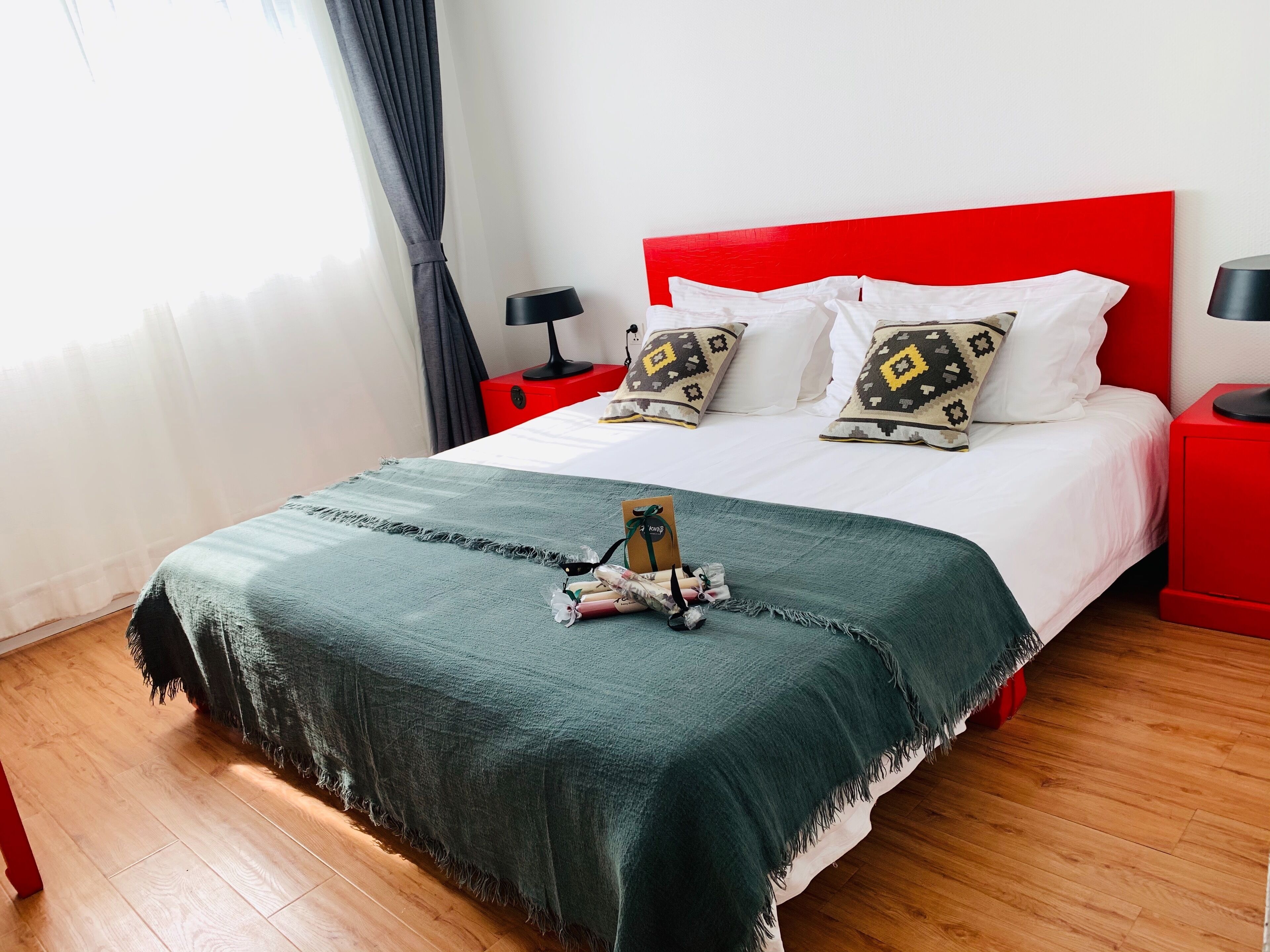 Una cama bien hecha con una manta verde, almohadas blancas y almohadas estampadas. Hay una mesita de noche roja con una lámpara y una pequeña mesa con una tarjeta y un regalo. La habitación tiene suelo de madera y una ventana con cortinas.
