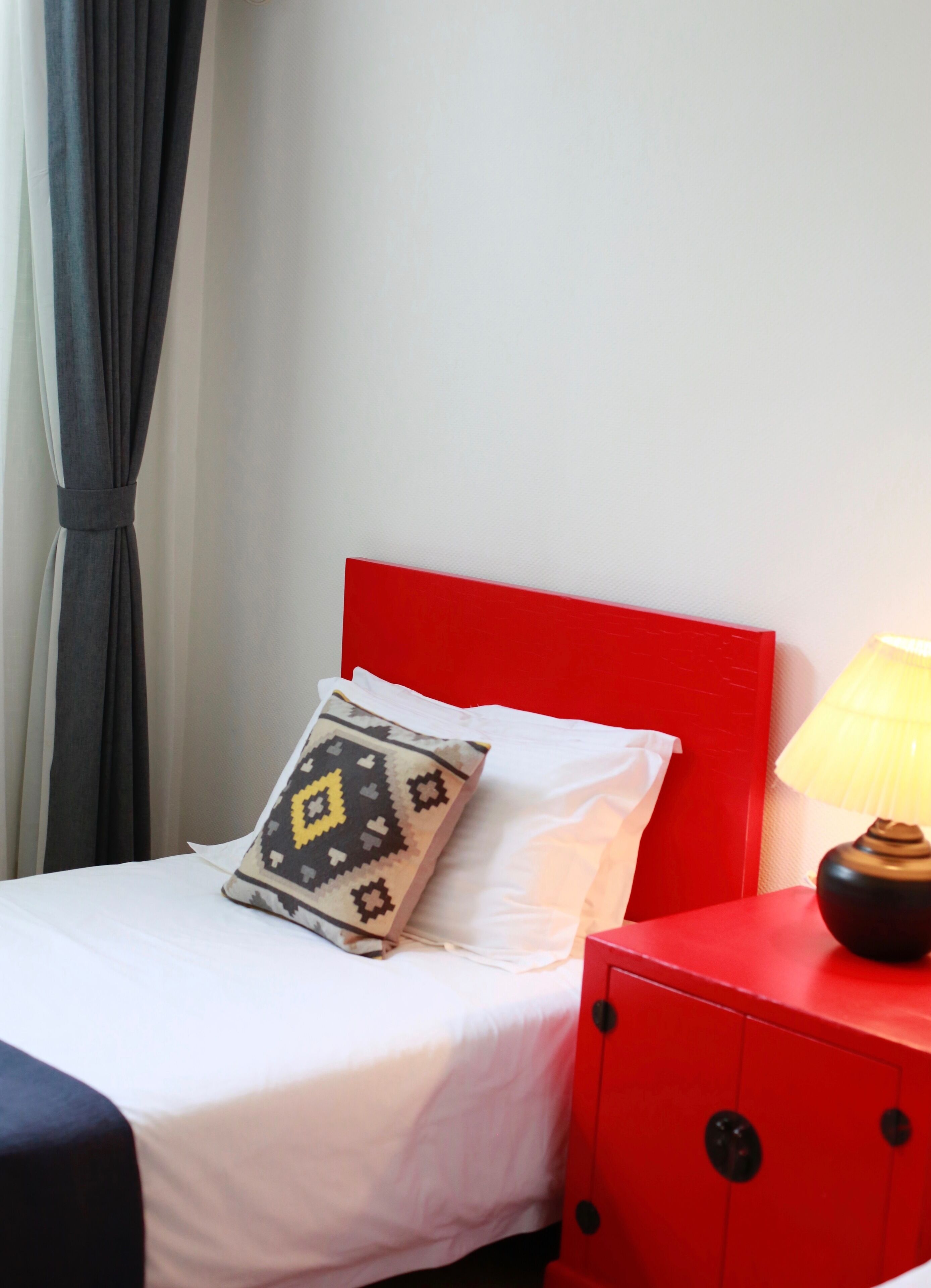 A bed with white bedding, a red headboard, and a patterned pillow. A red nightstand with a lamp is beside the bed.