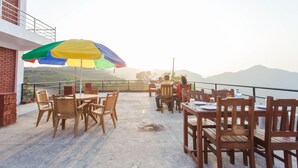 An outdoor patio with wooden tables and chairs, a colorful umbrella, and a view of the hills.