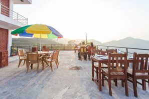 An outdoor patio with wooden tables and chairs, a colorful umbrella, and a view of the hills.
