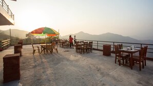 An outdoor dining area with tables and chairs, a colorful umbrella, and a view of mountains in the background.