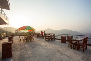 An outdoor dining area with tables and chairs, a colorful umbrella, and a view of mountains in the background.