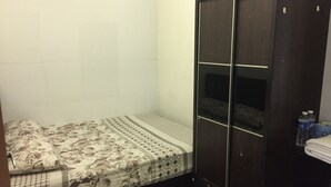 Een kleine, zwak verlichte kamer met een enkel bed, een donkere garderobe en een klein plankje met waterflesjes.