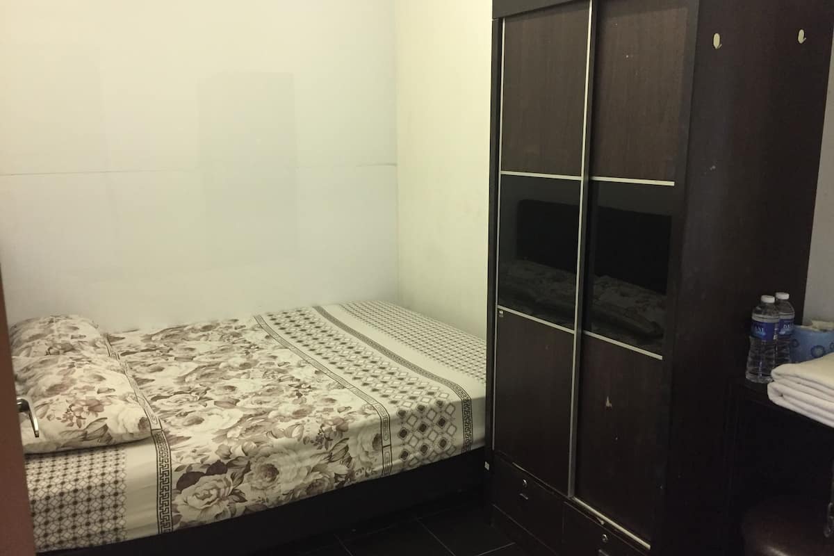 A small, dimly lit room with a single bed, a dark wardrobe, and a small shelf with water bottles.