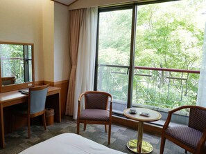 Habitación de hotel con cama, escritorio, dos sillas, una mesita con taza y plato, y un ventanal con vistas a árboles.