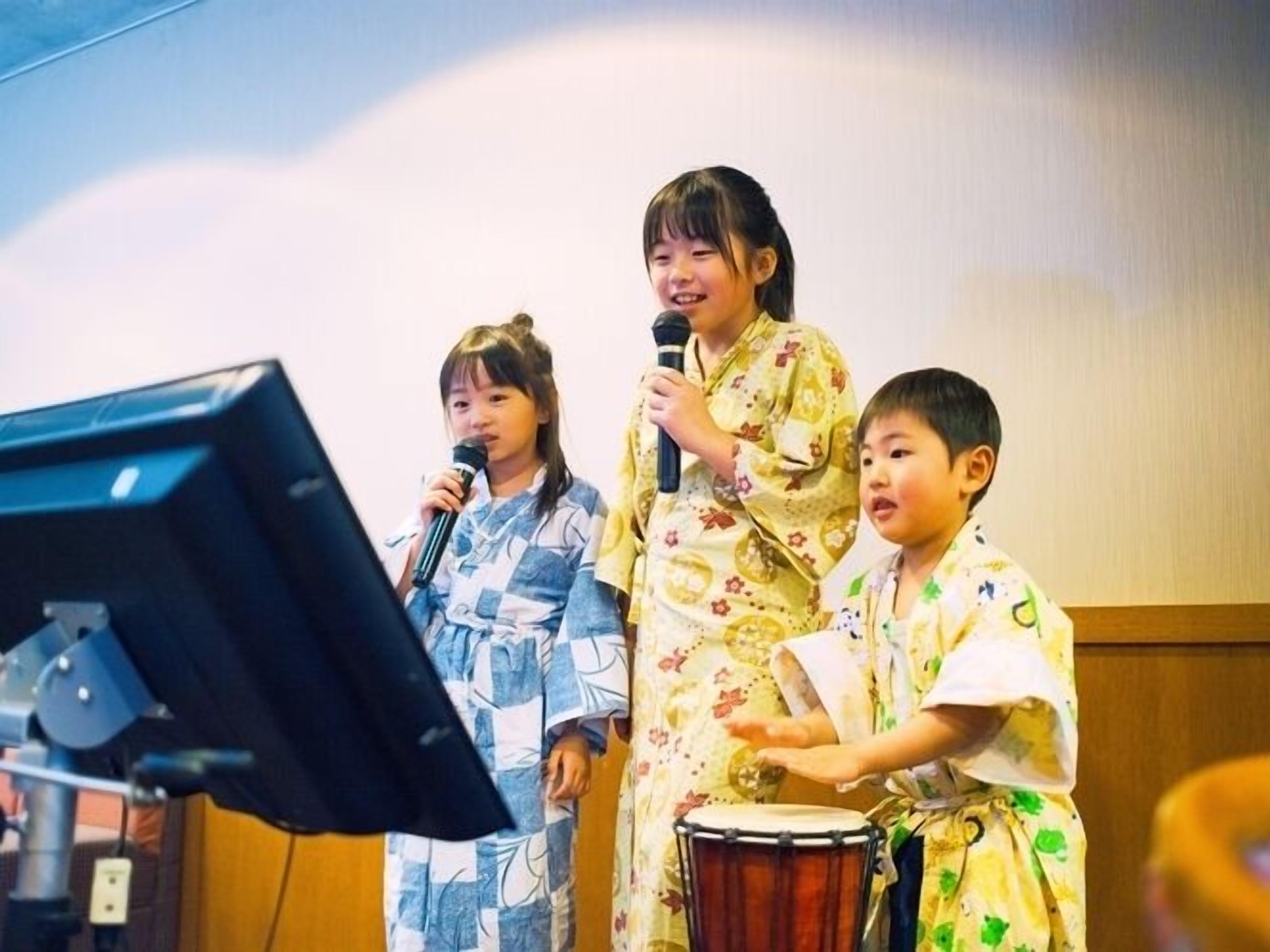 A group of children performing on stage with musical instruments and a microphone stand.