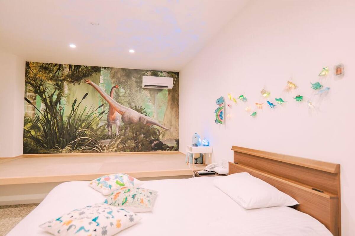 A bedroom with a mural of a dinosaur, a bed with white bedding, a nightstand, and a wall-mounted phone.