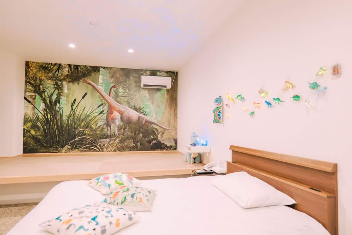 A bedroom with a mural of a dinosaur, a bed with white bedding, a nightstand, and a wall-mounted phone.