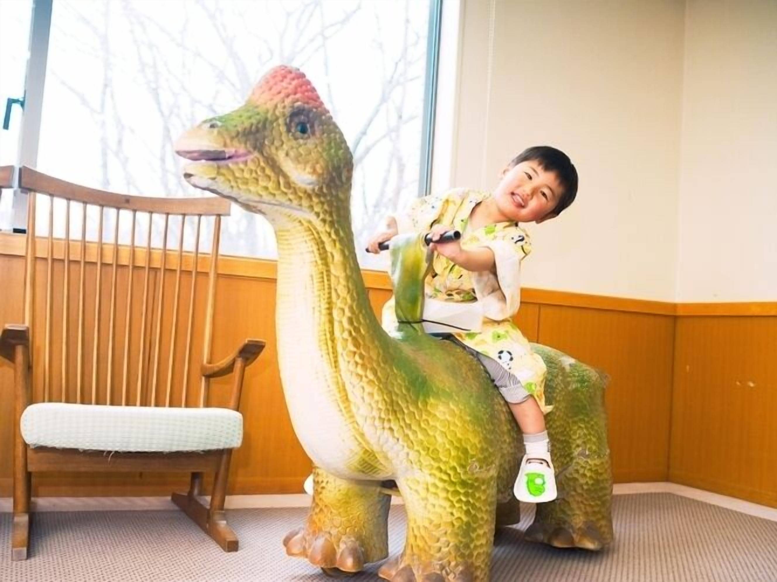 Un niño montando una silla con forma de dinosaurio en una habitación con muebles de madera y una ventana.