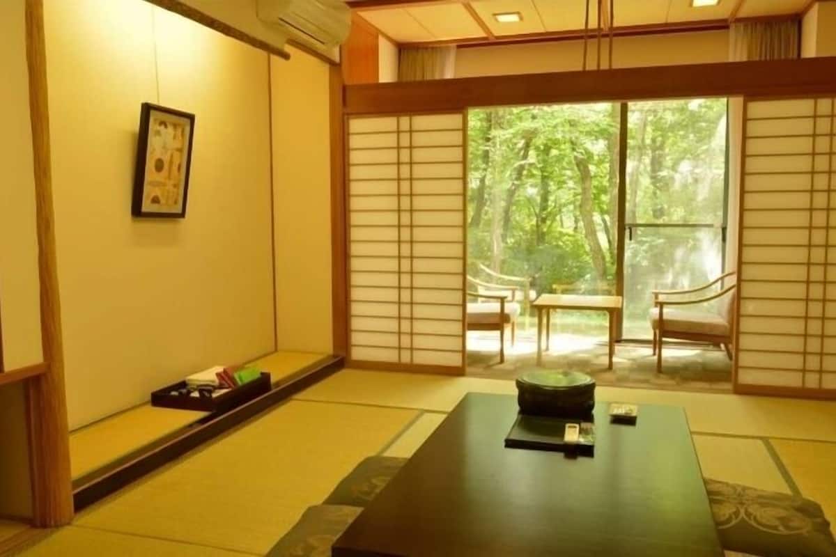 A traditional Japanese room with tatami flooring, a low table with a black tray, a framed picture on the wall, and a sliding door leading to a balcony with a view of trees.