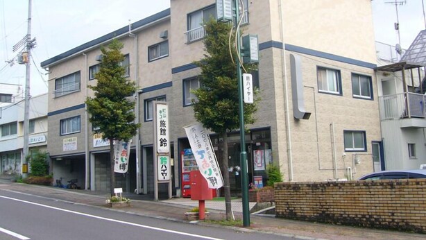 Um edifício de vários andares com fachada bege e detalhes azuis. Há várias placas em japonês, indicando diversos negócios ou serviços.