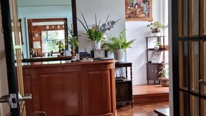 Un suelo de madera, una silla de madera, un armario de madera, una puerta de madera, un estante de madera, una mesa de madera, una planta en maceta, un reloj, un cuadro enmarco, una pared con un cuadro, un techo con una lámpara, y una ventana.