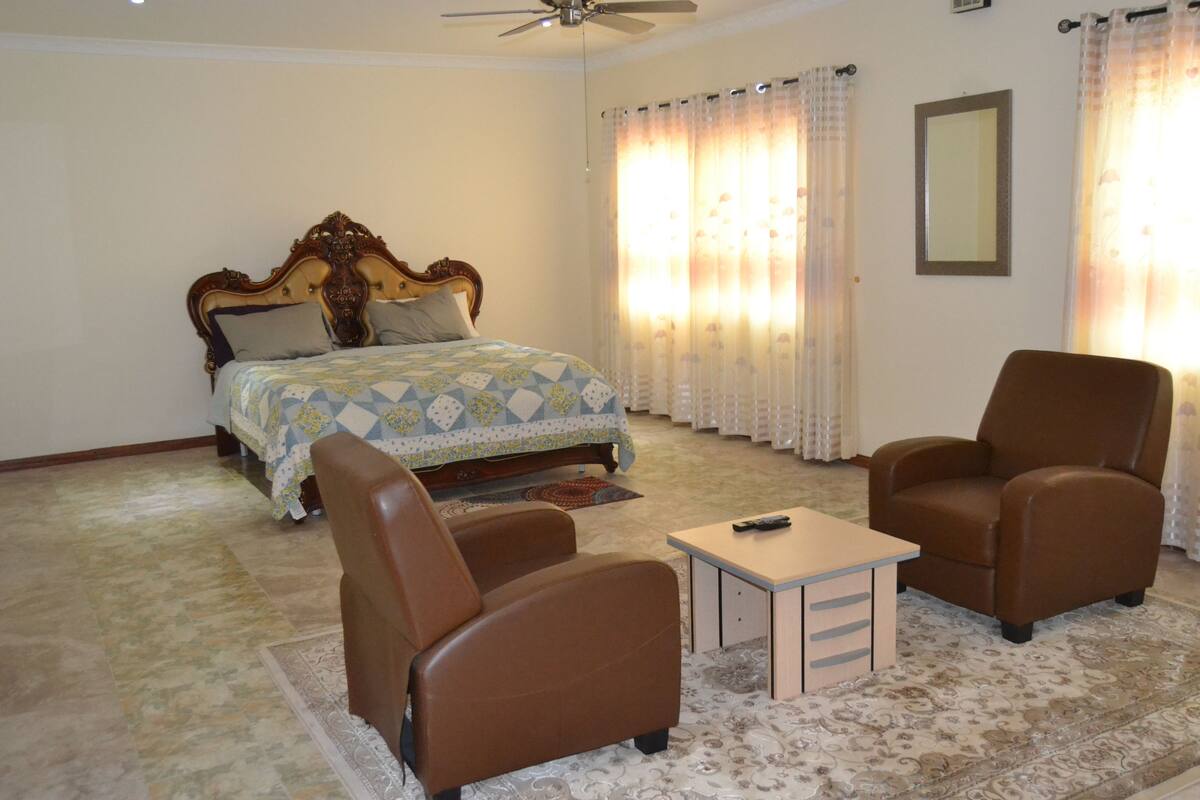A bedroom with a bed, two brown chairs, a small table, a ceiling fan, and a mirror on the wall.