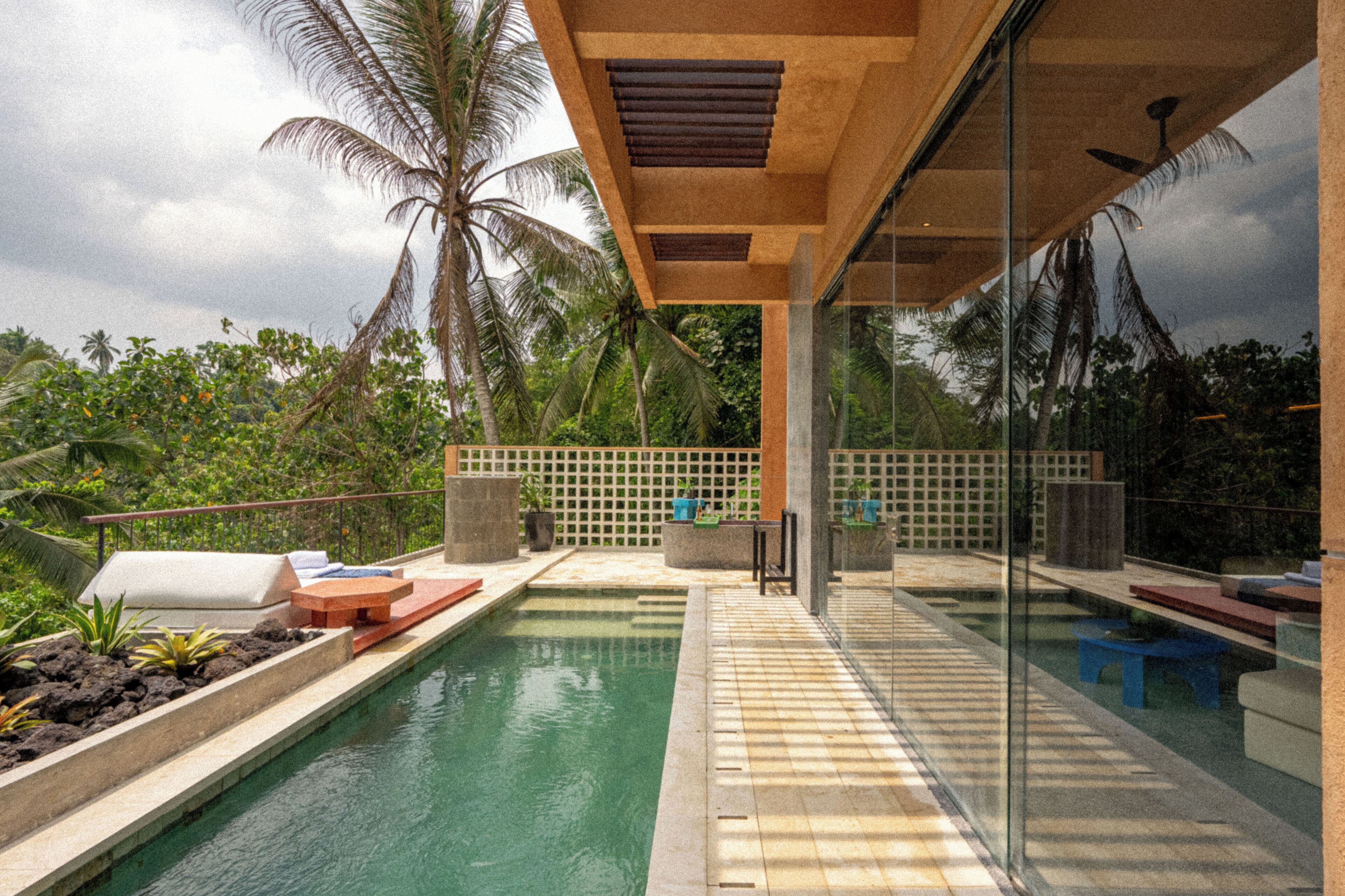 Un área de piscina moderna al aire libre con cerramiento de vidrio, palmeras y una zona de estar con cojines y una mesa.