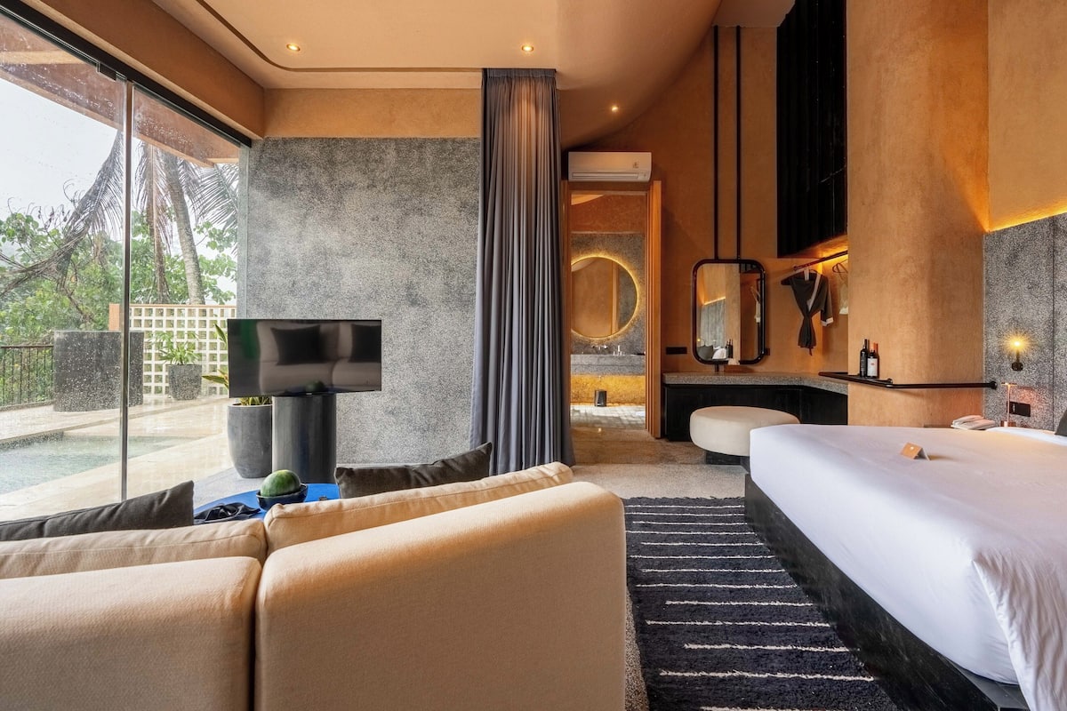 Una habitación de hotel moderna con una cama grande, un sofá, un televisor y un baño con espejo y lavamanos.