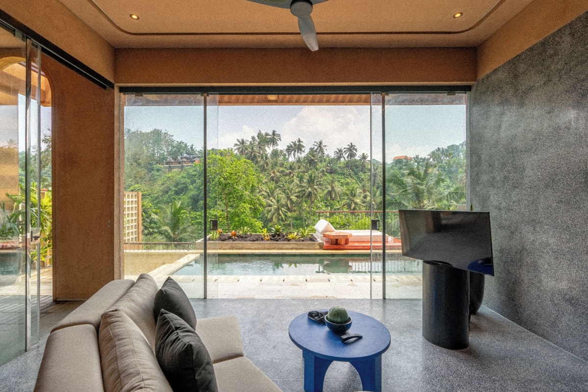 Un moderno salón con sofá, una mesita y televisor. Cuenta con ventanas amplias con vista a una piscina y palmeras.
