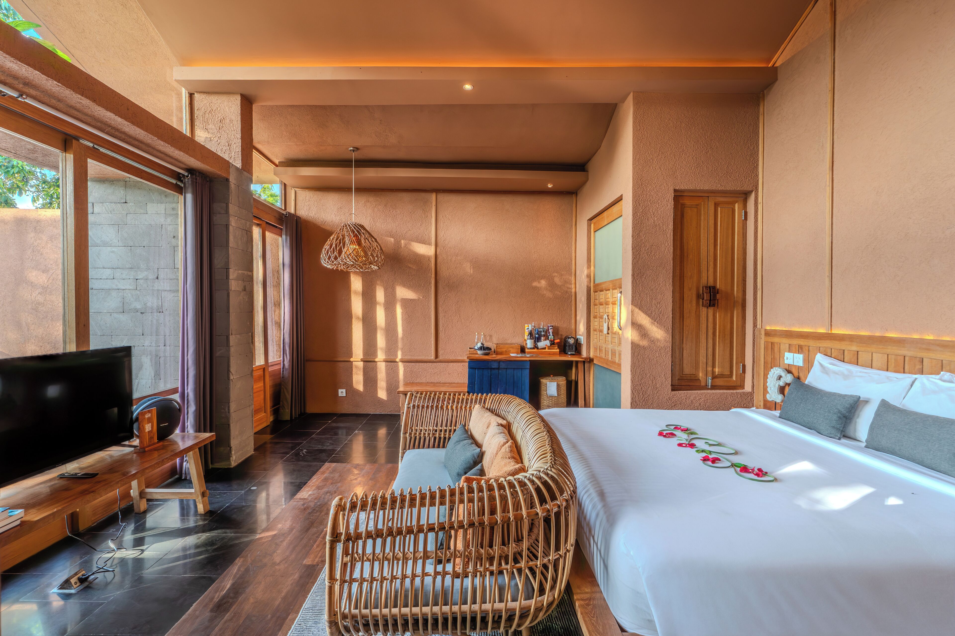 A modern hotel room with a large bed, a television, a wicker chair, and a small kitchenette.