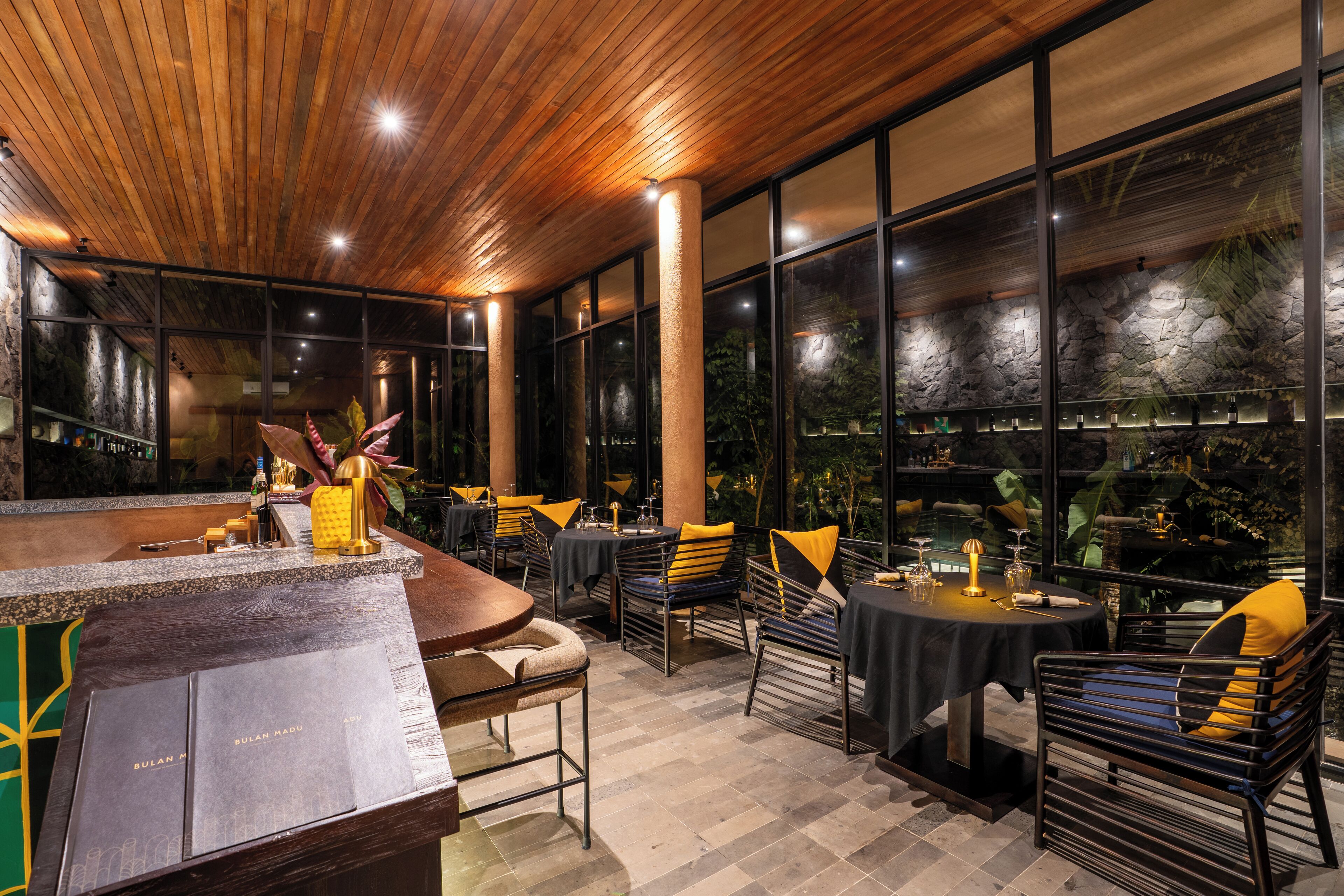 A restaurant with a wooden ceiling, large windows, and tables with yellow cushions.