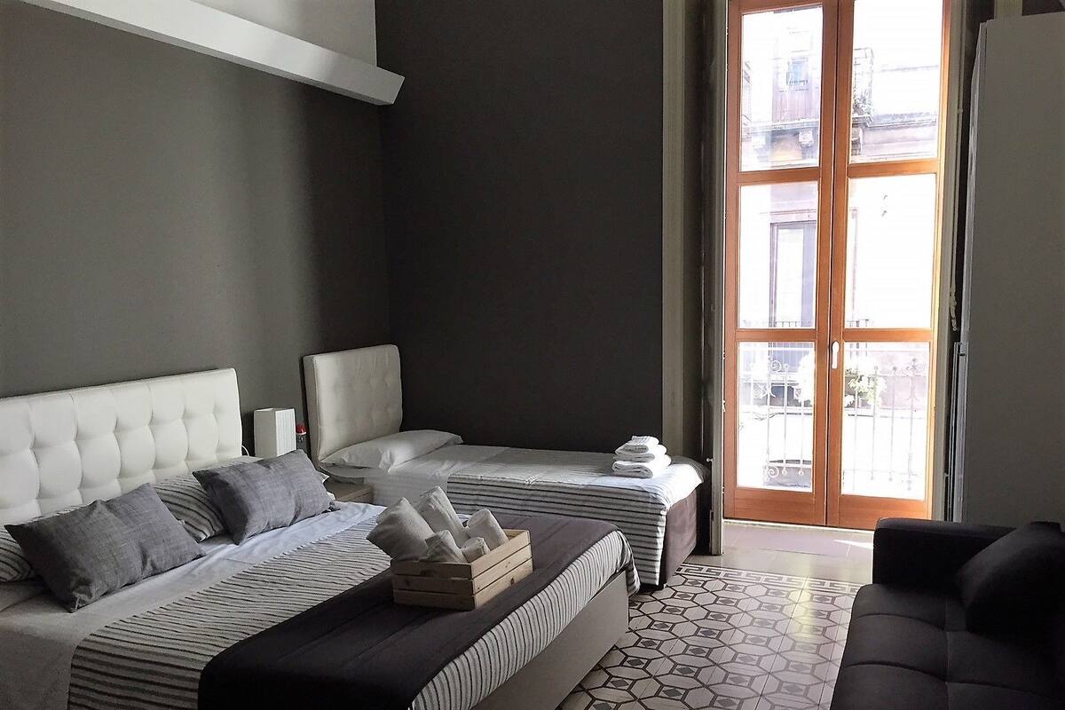 A hotel room with a large bed, a smaller bed, and a sofa. There is a window with a view of the outside.