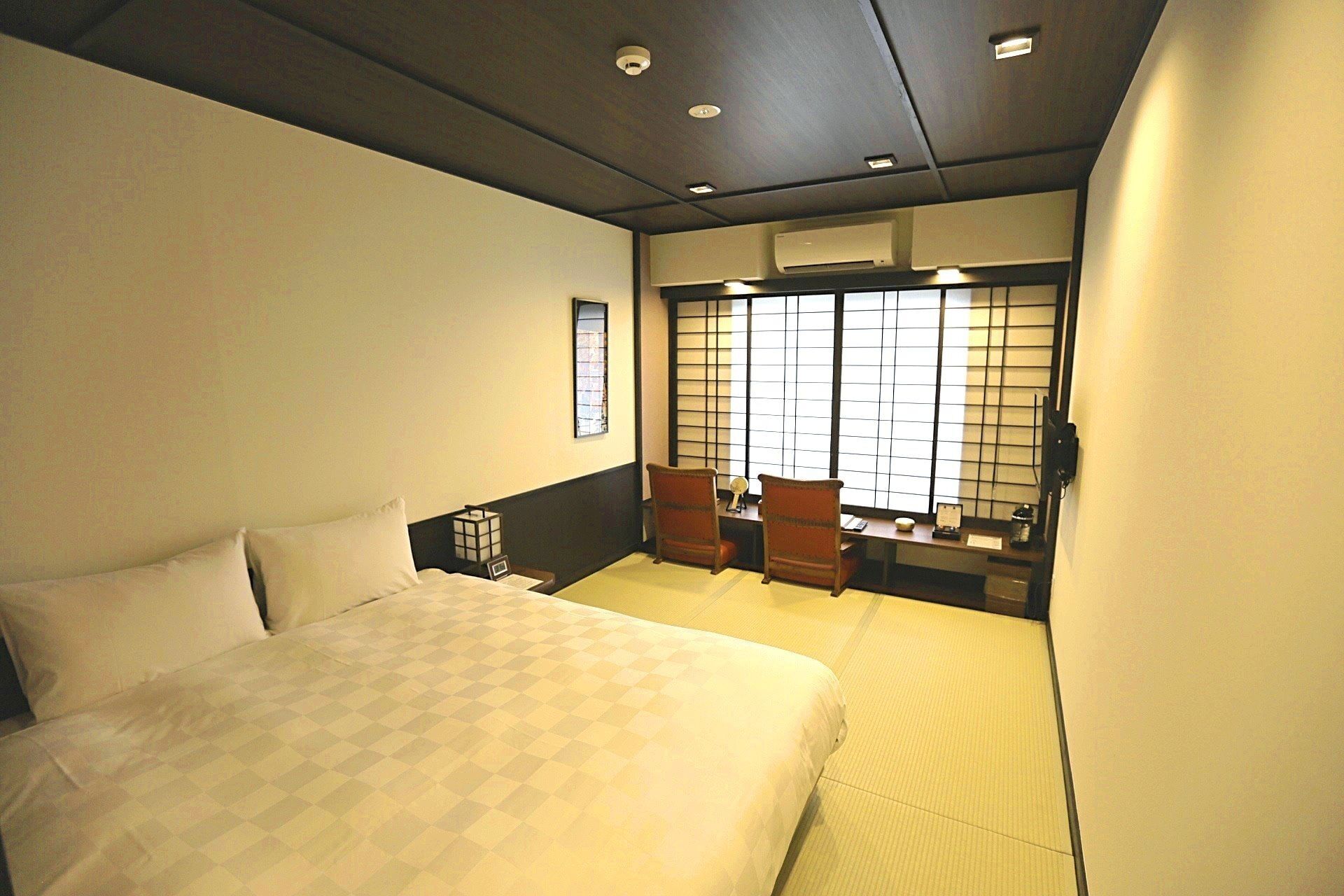 Quarto de hotel com uma cama grande, uma mesa com duas cadeiras, uma janela com persianas e um ar condicionado.
