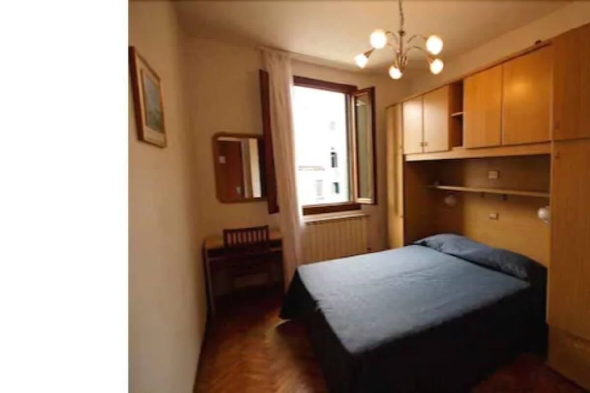 A narrow hallway leading to a bedroom with a bed, wooden wardrobe, and a radiator.