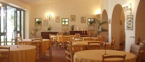 Un comedor con mesas y sillas redondas, cuadros en las paredes y un amplio ventanal que proporciona luz natural.