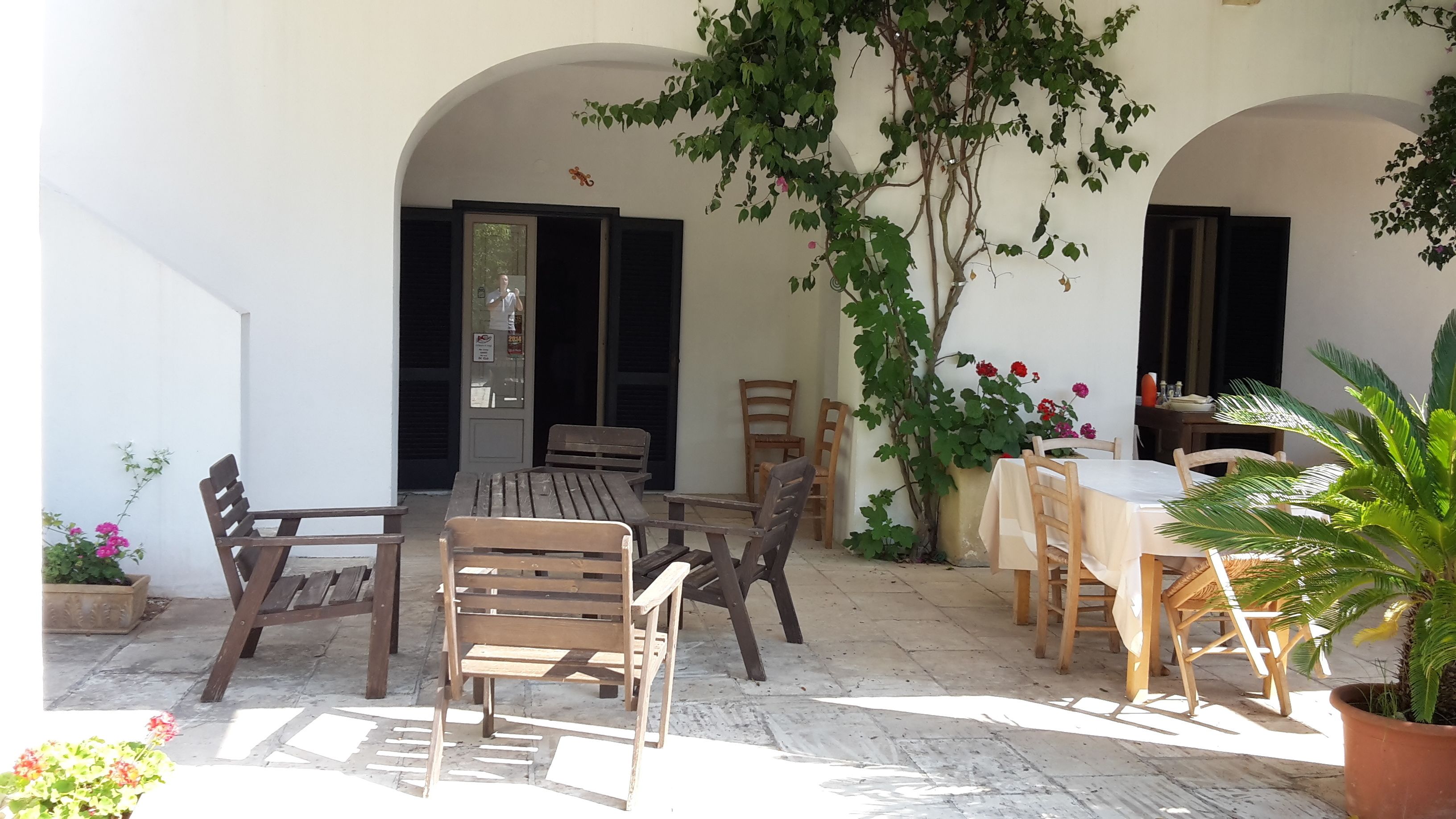 An outdoor patio with wooden furniture, including a table and chairs, surrounded by arches and potted plants.