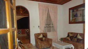A living room with a wooden door, a patterned armchair, a sofa with star-shaped cushions, a small table with a vase of flowers, and a window with curtains. - Tree House Holiday Home (Anuradhapura)