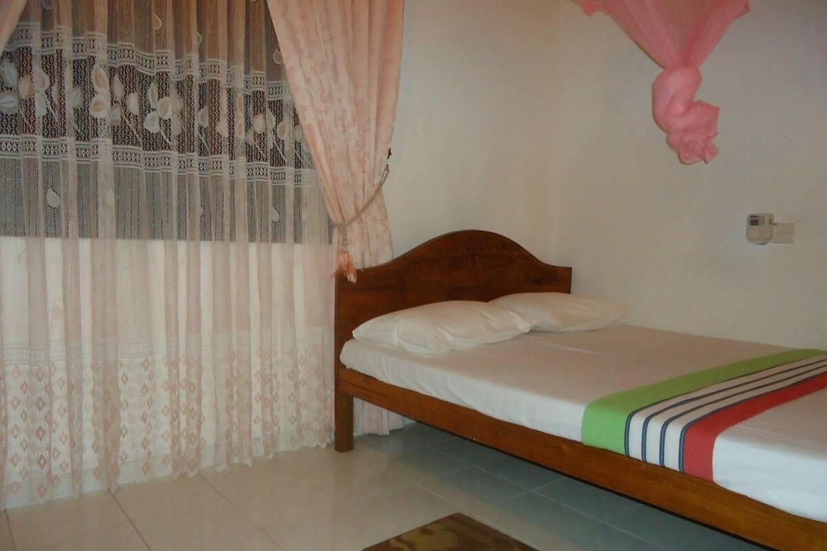 A single bed with a wooden frame, white bedding, and a colorful blanket. There is a window with sheer curtains and a pink decorative item hanging above the bed.