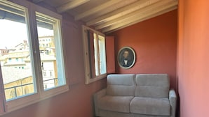 A room with a wooden floor, a grey sofa, and a window with a view of buildings. - Birbo Bike Hospitality (Siena)