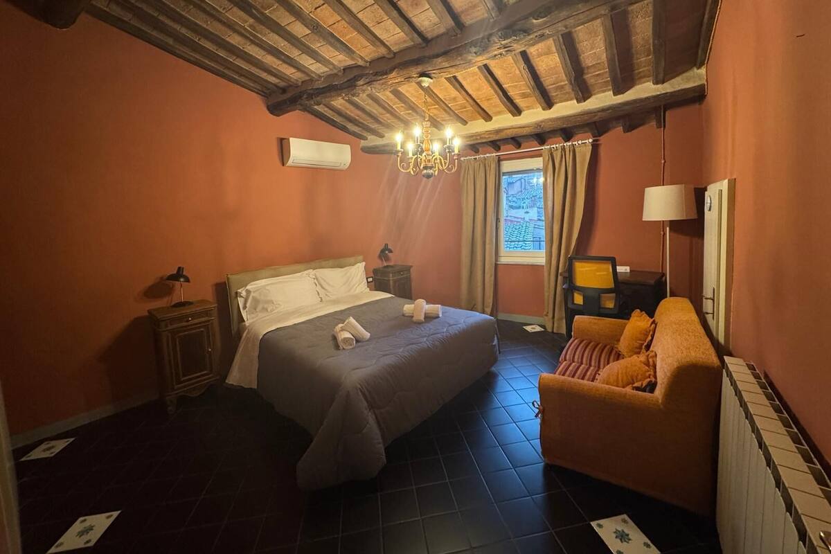 A bedroom with a bed, a sofa, a nightstand, an air conditioning unit, a chandelier, and a window with curtains.