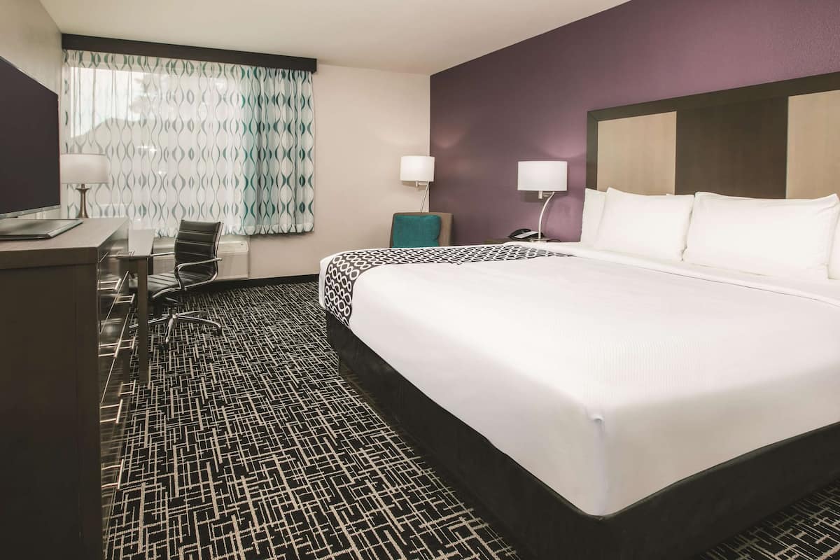 A hotel room with a large bed, a desk with a computer, a chair, and a patterned carpet.