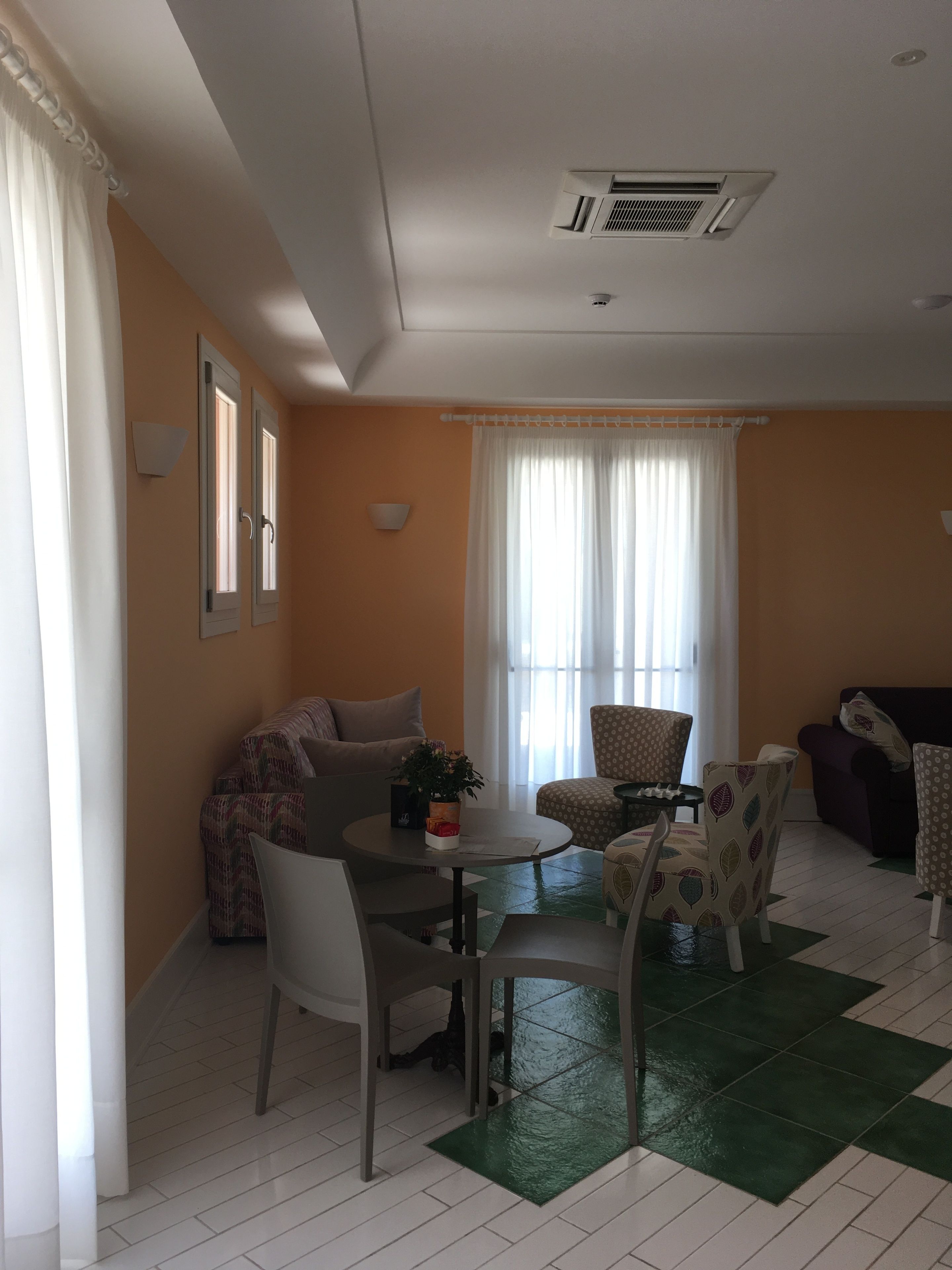 Una sala de estar con un sofá, sillas, una mesa de centro y una mesa de comedor. Hay dos ventanas con cortinas, una rejilla de ventilación en el techo y una lámpara empotrada en la pared.