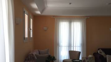 Una sala de estar con un sofá, sillas, una mesa de centro y una mesa de comedor. Hay dos ventanas con cortinas, una rejilla de ventilación en el techo y una lámpara empotrada en la pared.