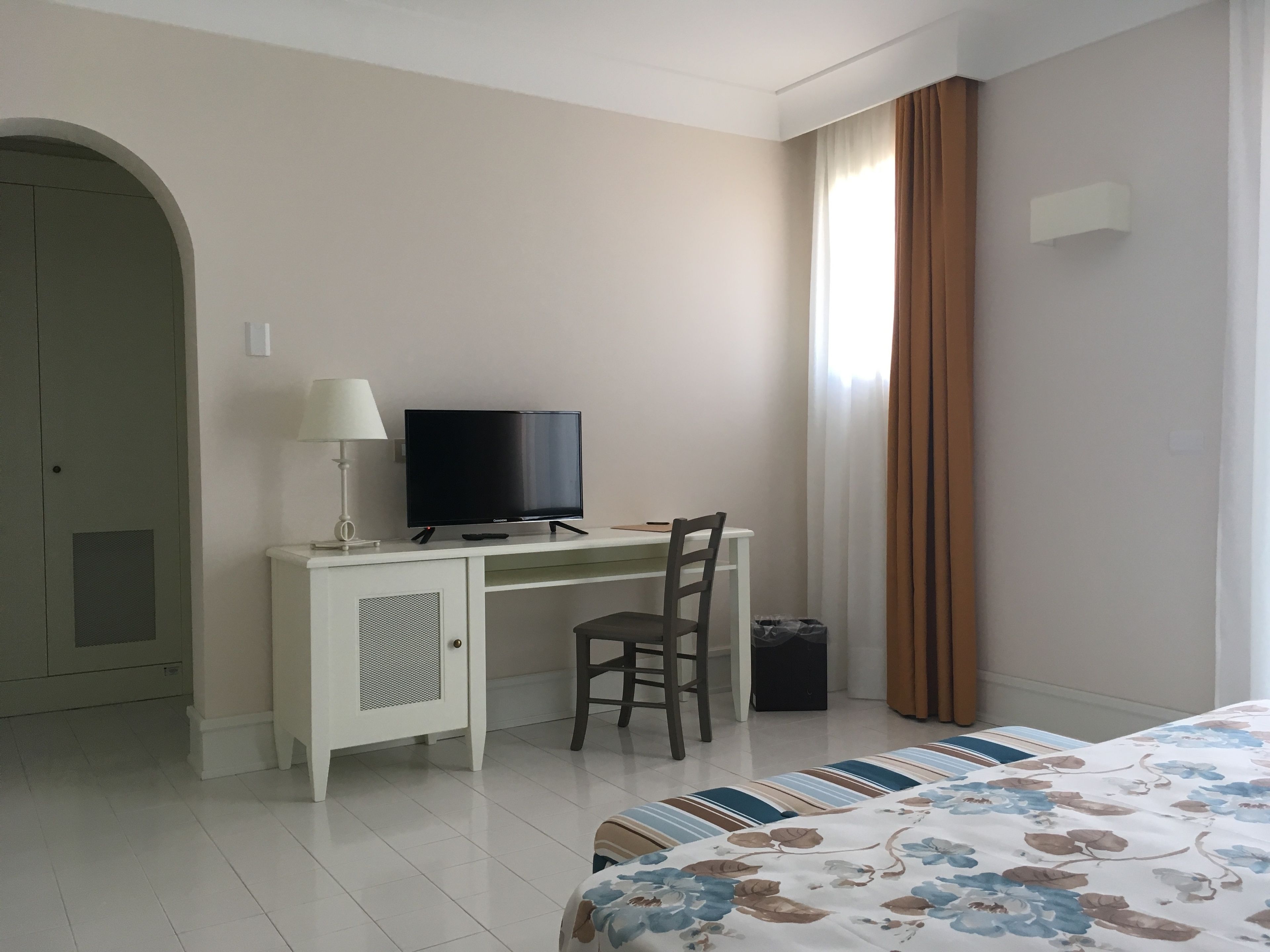 Habitación de hotel con cama, escritorio con televisor, una silla, lámpara y ventana con cortinas.