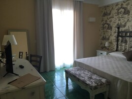 Habitación de hotel con cama, escritorio con lámpara, silla, banco y ventana con cortinas.