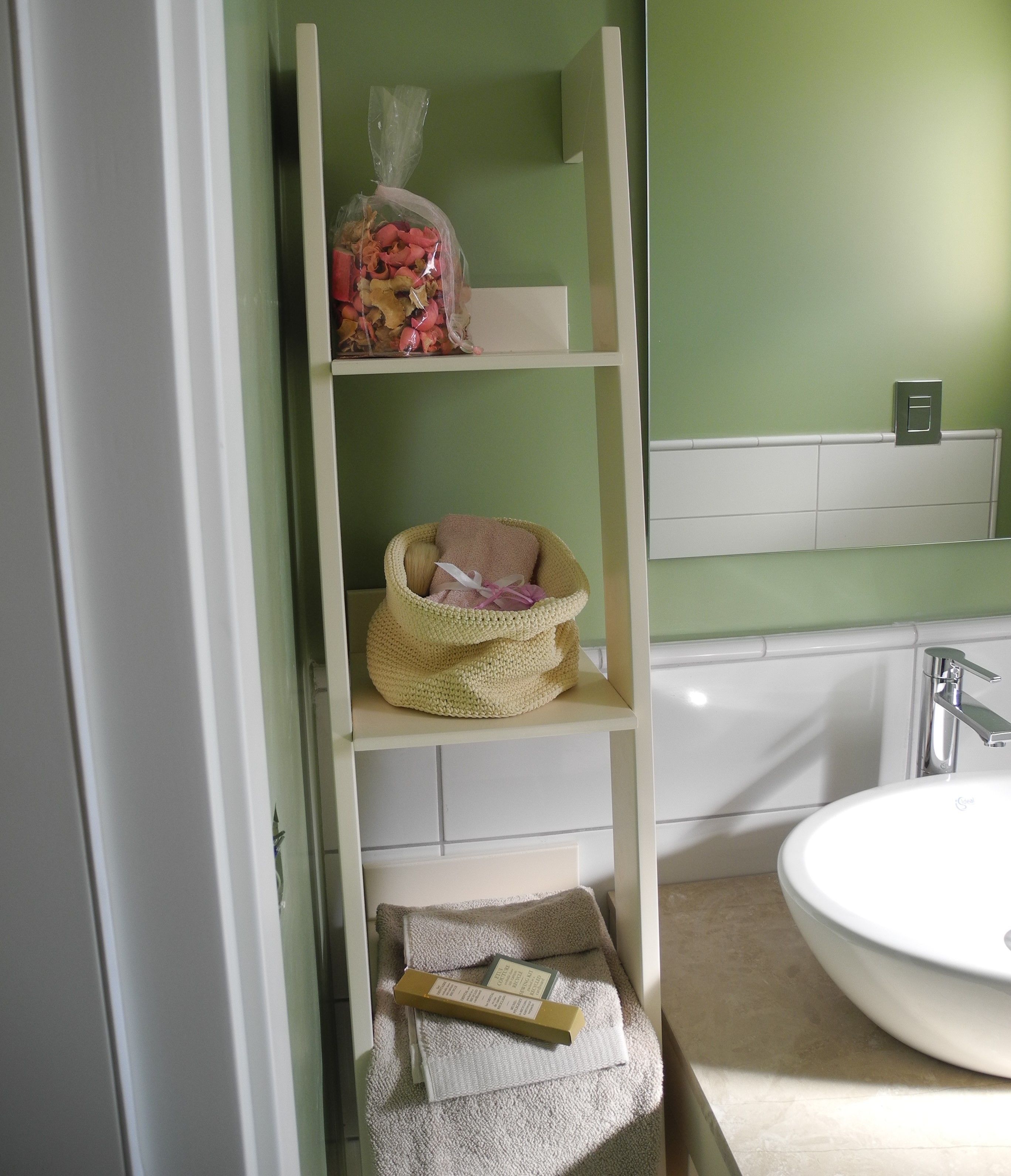 Un bagno con un lavandino bianco, pareti verdi e uno scaffale bianco con un cesto e una borsa.