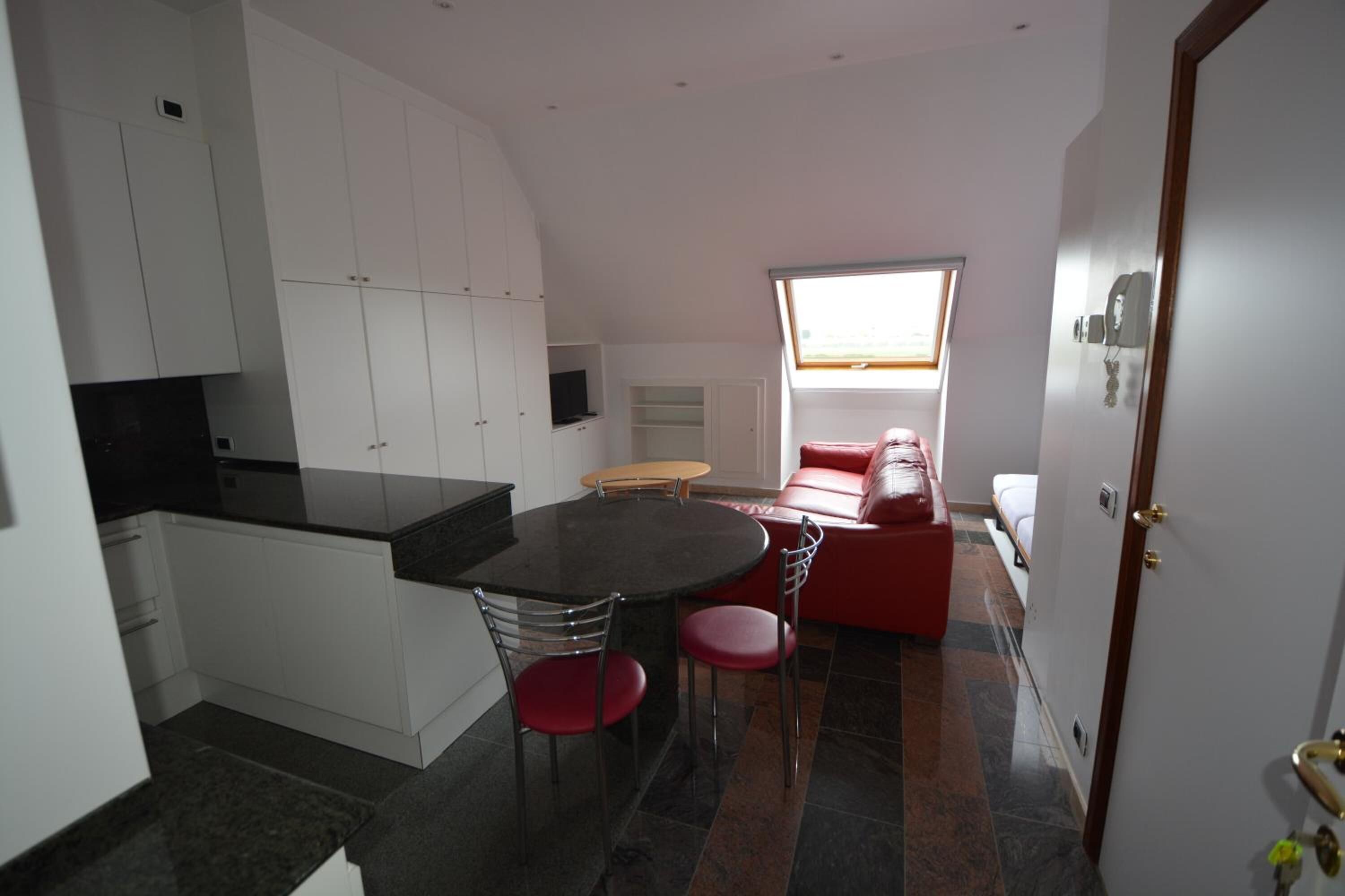 Een kleine keuken met witte kastjes, een zwart aanrecht en een eethoek met een ronde tafel en rode stoelen.