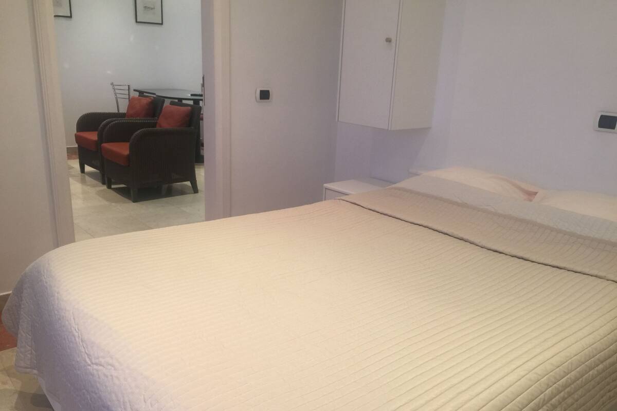 Een slaapkamer met een bed, een nachtkastje, een kledingkast, en een stoel in de aangrenzende kamer.