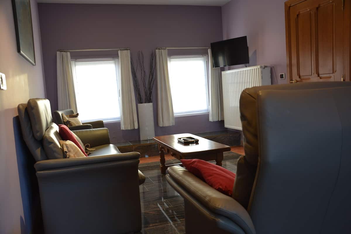 Een woonkamer met een bank, fauteuil en salontafel. Er zijn twee ramen met gordijnen, een radiator en een televisie aan de muur.