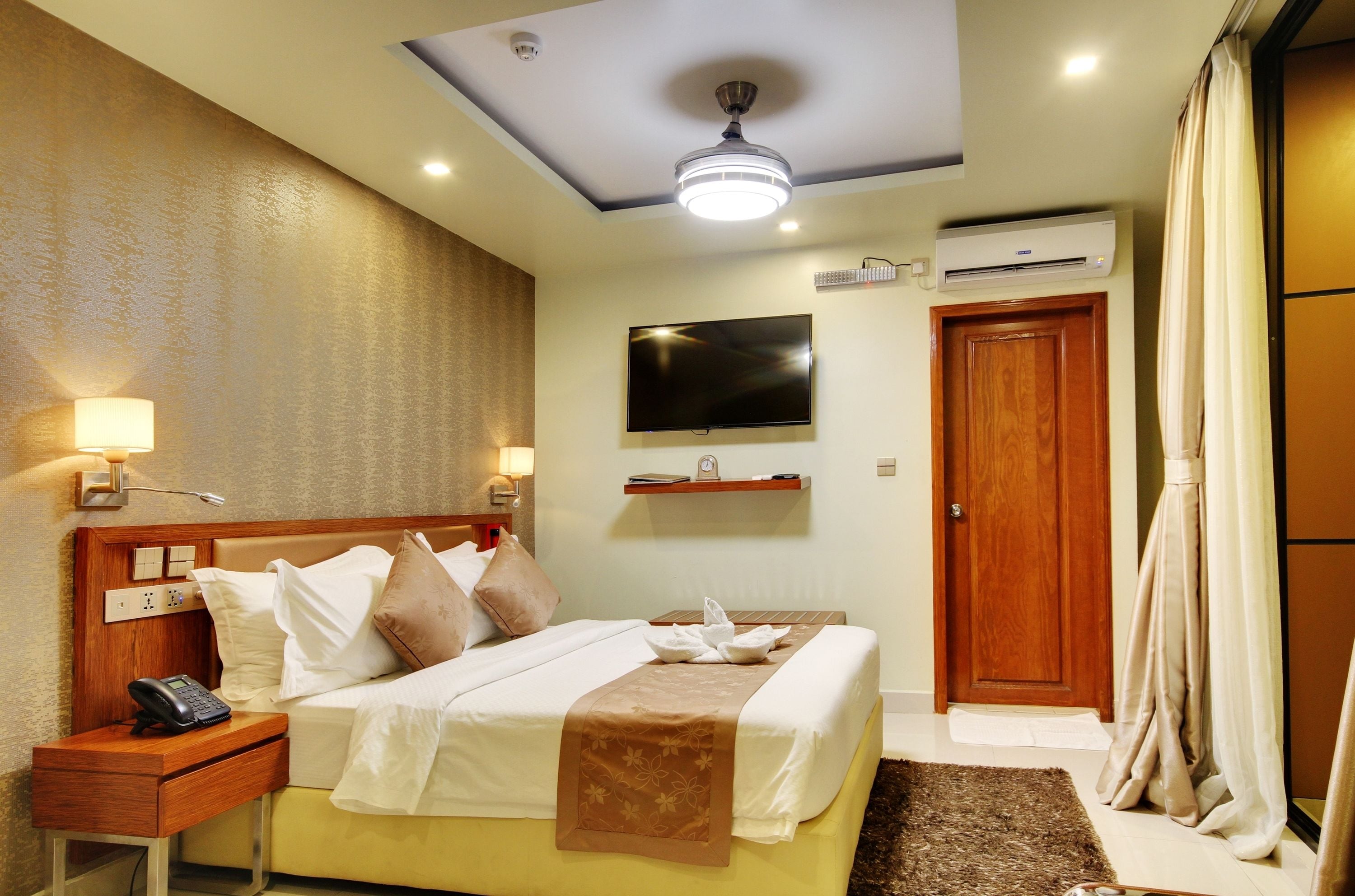 A hotel room with a bed, bedside table with a phone, a flat-screen TV mounted on the wall, an air conditioning unit, a wooden door, and a curtain.
