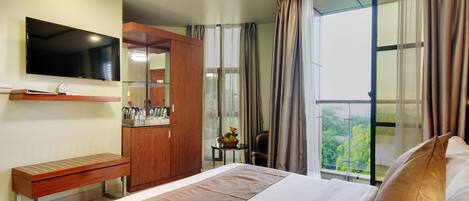 A hotel room with a bed, a TV mounted on the wall, a wooden bench, a small table with a fruit bowl, and a view of greenery through the window.