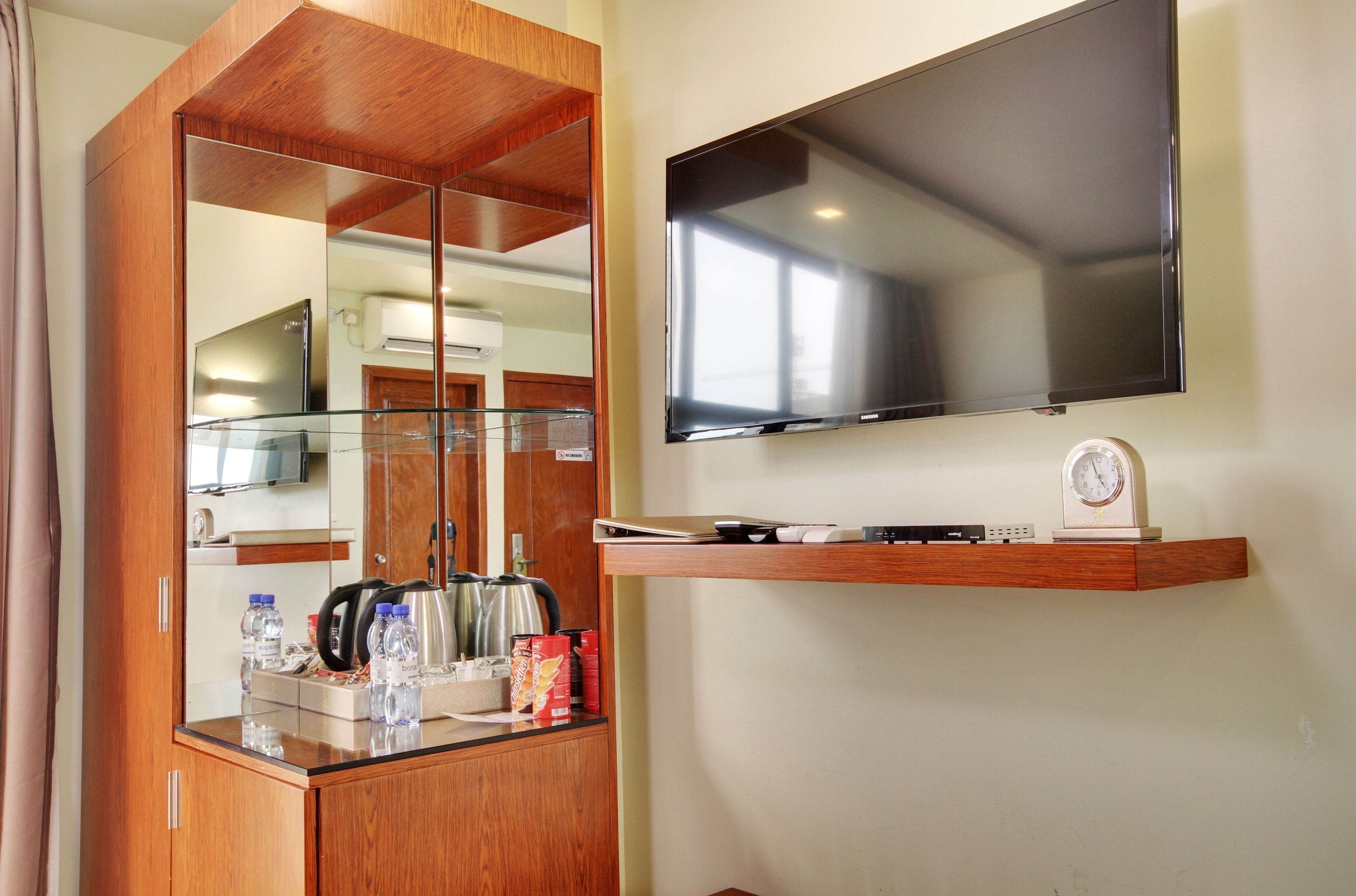 A hotel room with a wooden wardrobe, a glass-top minibar, a flat-screen TV, and a small shelf with a clock and a remote control.