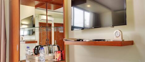 A hotel room with a wooden wardrobe, a glass-top minibar, a flat-screen TV, and a small shelf with a clock and a remote control.