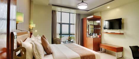 Habitación de hotel con cama, mesita de noche, lámpara, televisor, escritorio, silla y ventana con cortinas.