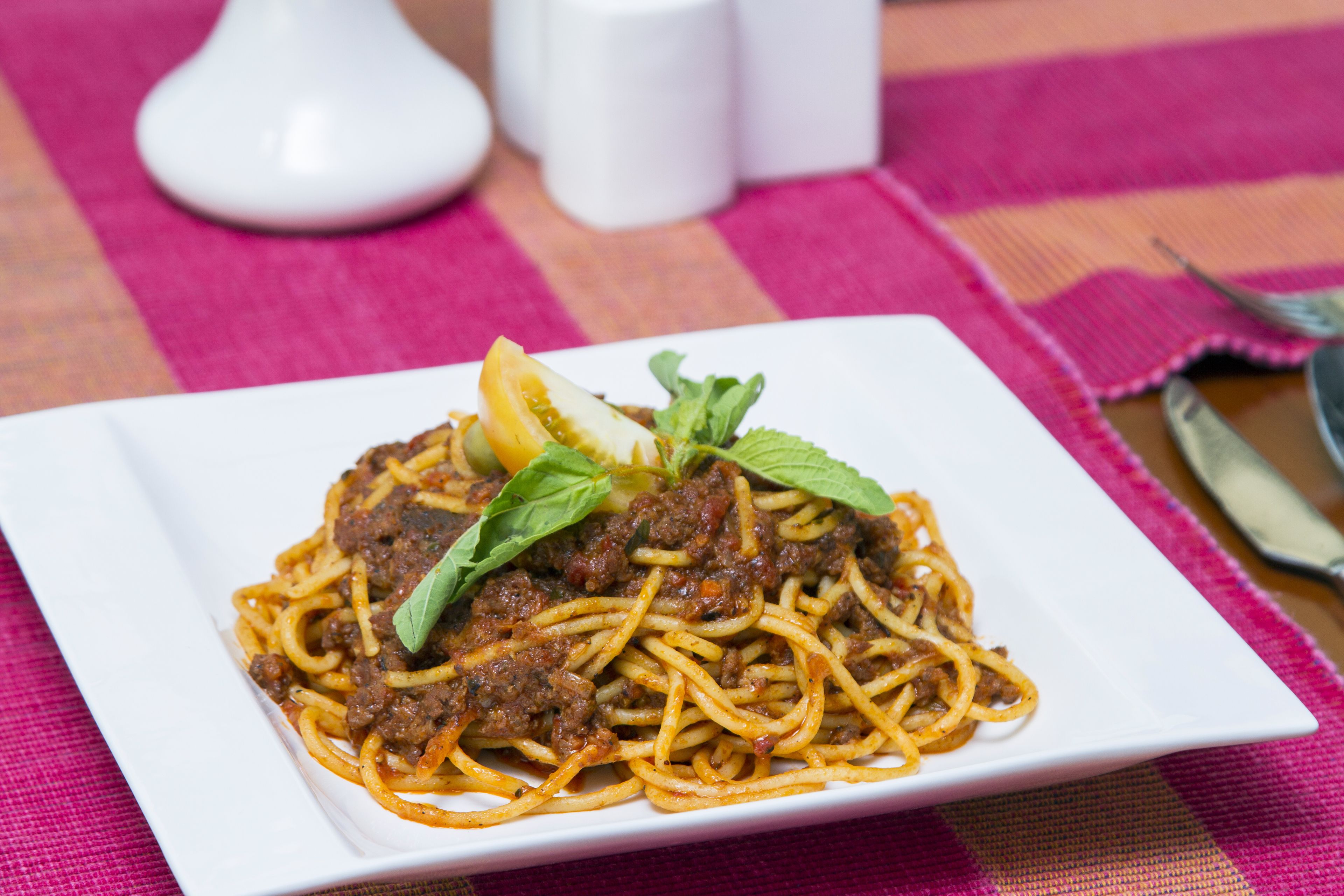 Een bord spaghetti met vleessaus, versierd met een citroenpartje en muntblaadjes, op een wit bord.